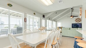 A dining area with a white table and chairs, a green sofa, and a bar area with stools. - 2nd Row Gulf View Home w/ 2 Decks, Board Games, 3 Kings+Bunk all w/Deck Access (Galveston)