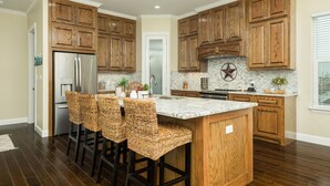 Una cocina con gabinetes de madera, un refrigerador de acero inoxidable, encimera de mármol y una zona de bar con sillas de mimbre.