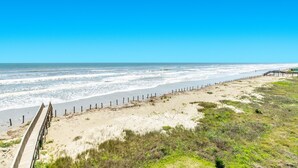 Una playa con un paseo marítimo de madera que conduce al agua, dunas arenosas y un cielo azul despejado.