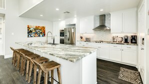 A modern kitchen with white cabinets, a large island with stools, stainless steel appliances, and a colorful abstract painting on the wall. - Private Canal Home w/ 2 Living Areas, Easy Bay Access & POOL (Galveston)