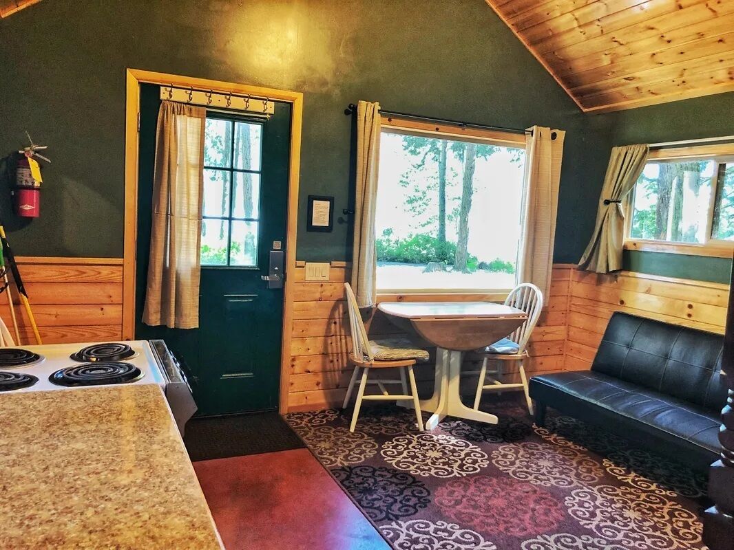 A cozy cabin interior with a dining table, chairs, a sofa, a stove, and a window with curtains.