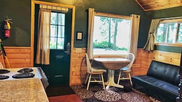 A cozy cabin interior with a dining table, chairs, a sofa, a stove, and a window with curtains.