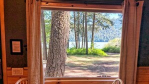 Une cabane en bois avec une table et des chaises, une fenêtre avec des rideaux, et une vue sur une forêt et un lac.