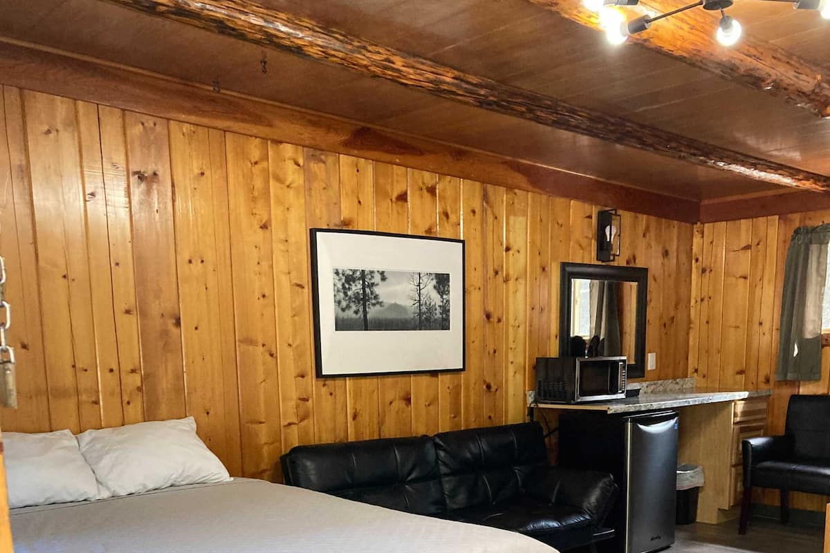 Um quarto com painéis de madeira, uma cama, um sofá de couro preto, uma pequena cozinha compacta e uma escrivaninha.