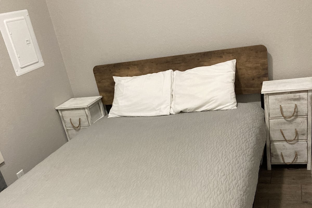 Uma cama bem arrumada, com estrutura de madeira e um espalhão cinza texturizado. Há duas mesas de cabeceira com gavetas brancas e uma pequena mesa lateral de madeira.