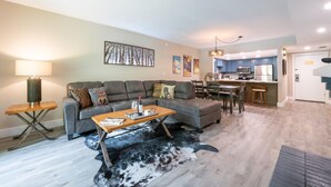 Ein modernes Wohnzimmer mit einer Couch, einem Couchtisch und einem Teppich. Im Hintergrund ist eine Küchenzeile mit Esstisch und Stühlen zu sehen.