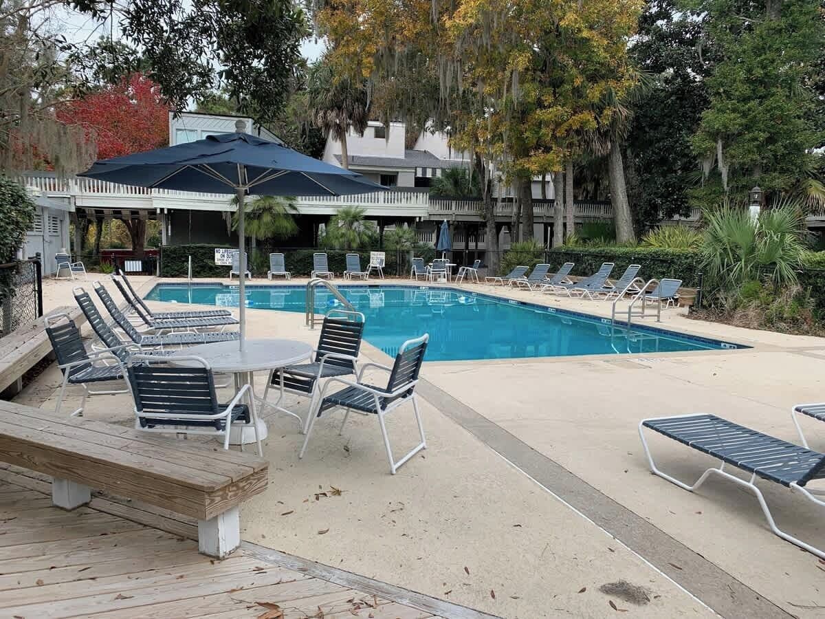 Un espace piscine avec des chaises longues et une table, entouré d’arbres et d’un bâtiment délimité par une clôture blanche.