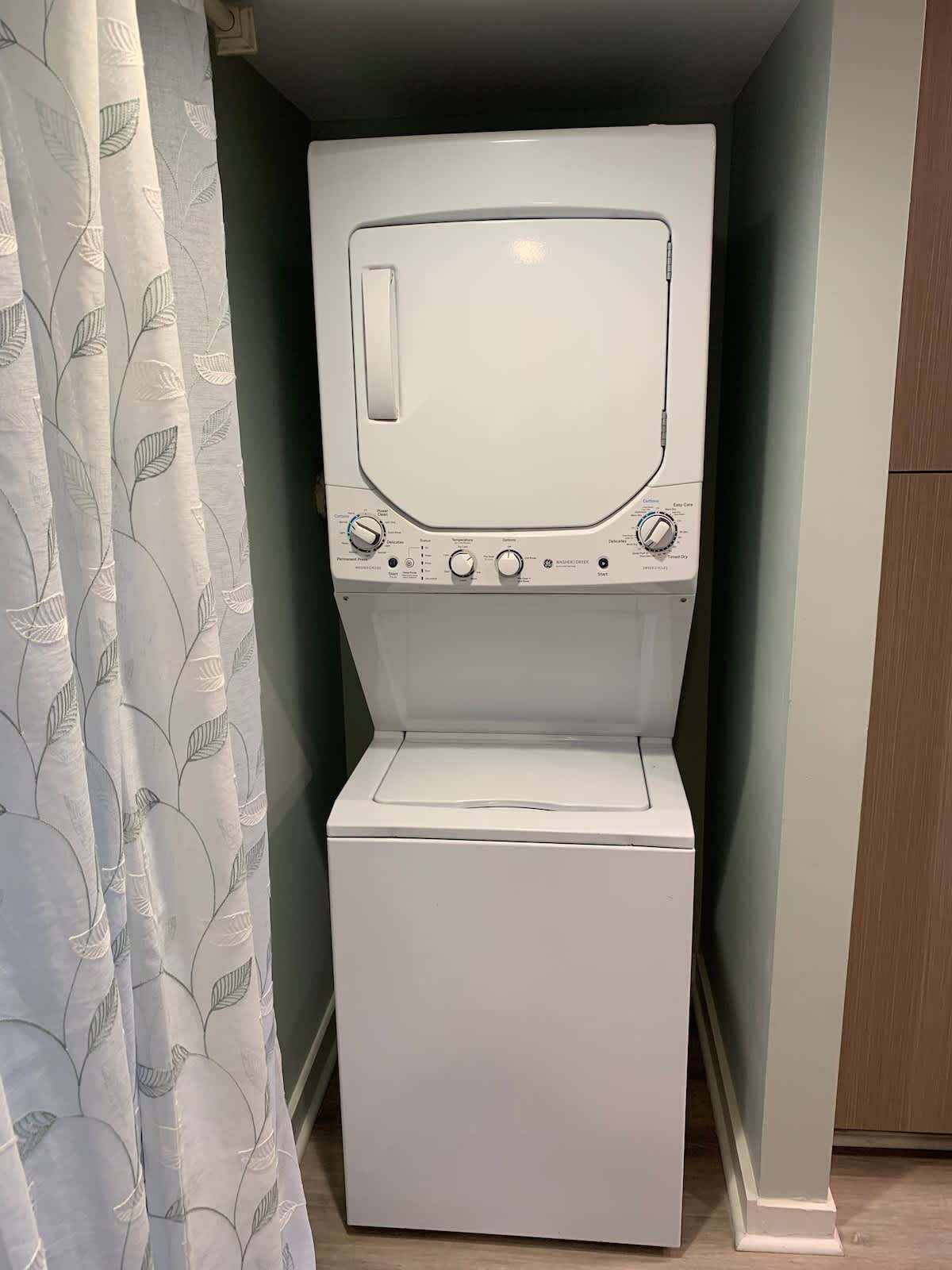 A compact, white washing machine in a small laundry room with a leaf-patterned shower curtain.