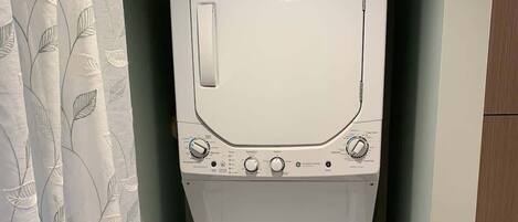 A compact, white washing machine in a small laundry room with a leaf-patterned shower curtain.