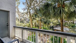 A balcony with a white railing, a chair, and a view of a garden with various trees and a small bridge. - Poolside Beach Villa 2 bed 2 & one half bath-Pet Friendly (Hilton Head Island)