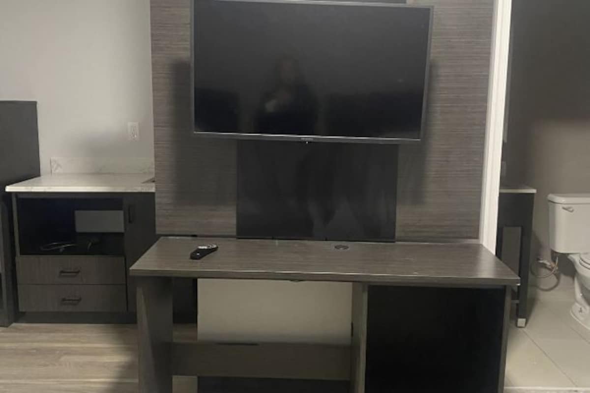 A modern living room with a flat-screen TV mounted on a wooden wall, a small wooden table with a remote control, and a white toilet in the background.