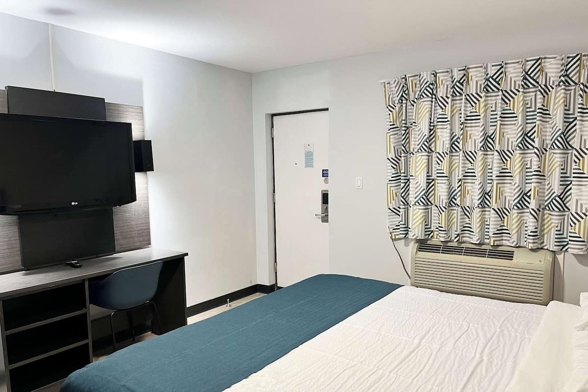 A hotel room with a bed, a desk with a flat-screen TV, a chair, and a patterned curtain.