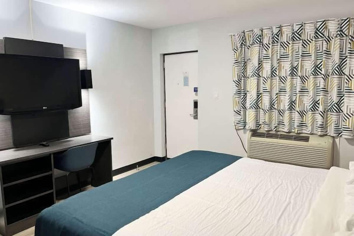 A hotel room with a bed, a desk with a flat-screen TV, a chair, a patterned curtain, and a wall-mounted air conditioner.