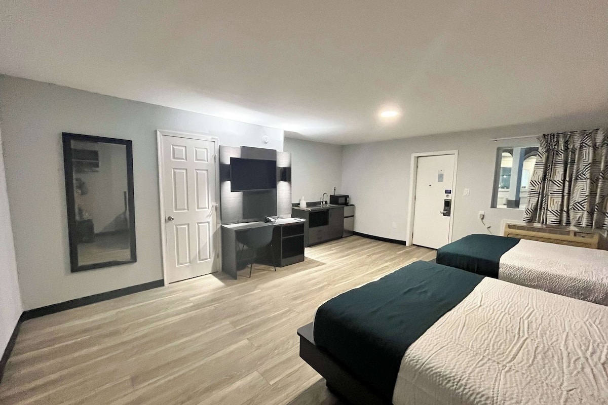 A hotel room with a bed, a desk with a chair, a TV, and a small kitchenette.