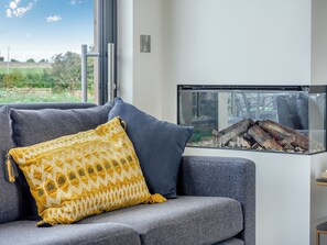 A modern living room with a grey sofa, a yellow patterned cushion, and a glass fireplace with logs. - Buttercup Lodge - UK36655 (Boothby Pagnell)