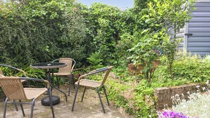 Een terras buiten met rieten stoelen en een ronde tafel, omgeven door weelderige begroeiing en bloeiende planten.