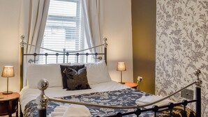 Een net opgemaakt bed met een metalen frame, witte beddengoed en een bloemenprint deken. Twee bedlampen staan op houten nachtkastjes, en er is een raam met gordijnen.