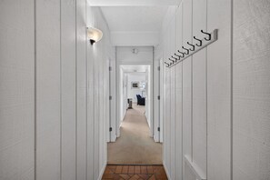 Un couloir aux murs blancs, avec un luminaire, un porte-manteau et un sol recouvert d’une moquette, qui mène à une pièce contenant une chaise et une lampe.