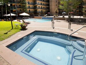 Una piscina rettangolare con bordo piastrellato, circondata da un solarium in mattoni. Sullo sfondo, lettini, ombrelloni e un edificio a più piani con balconi.