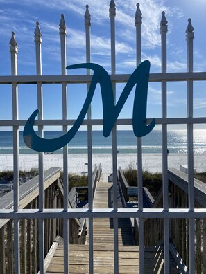 Una passerella in legno conduce a una spiaggia con un recinto bianco con un grande logo blu 'M'.