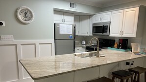 Una cocina con armarios blancos, un refrigerador de acero inoxidable, un microondas, un lavamanos y taburetes de bar de madera.