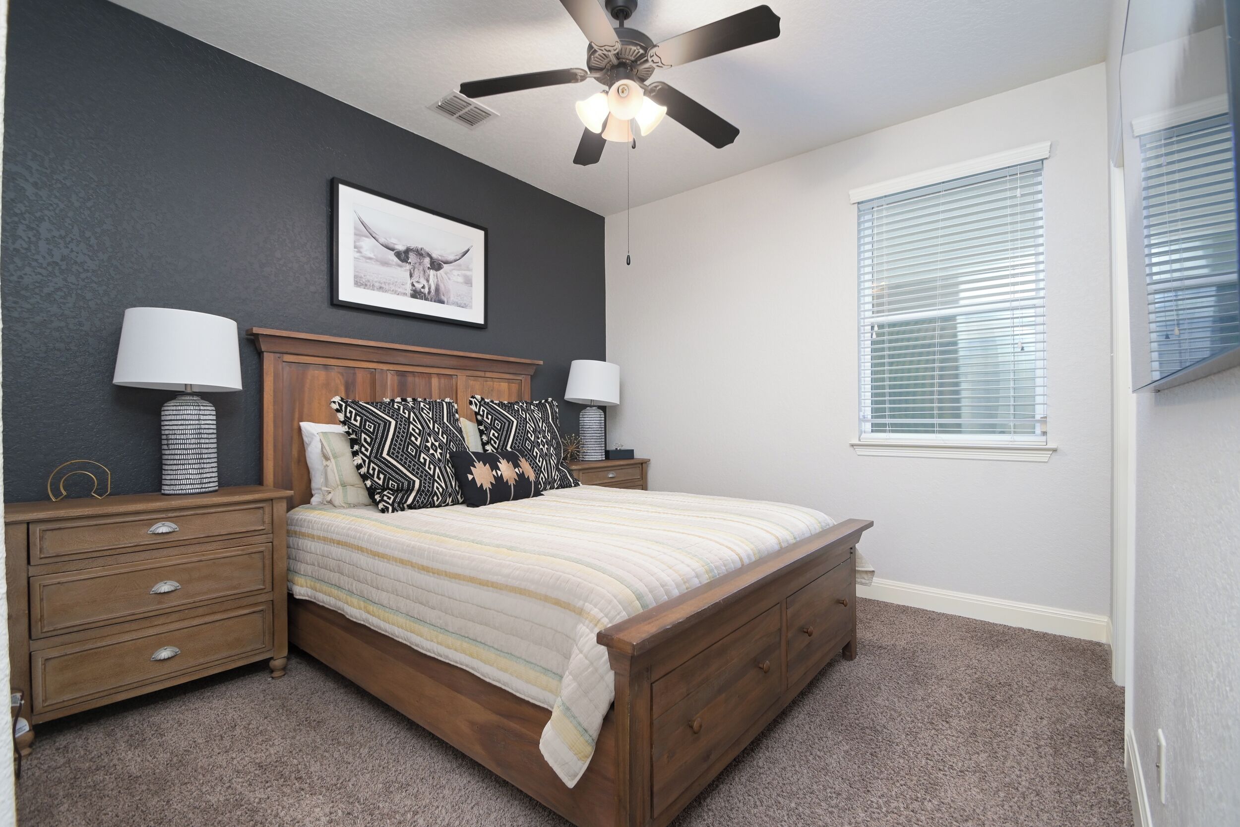 A bedroom with a wooden bed, two bedside tables with lamps, a ceiling fan, a framed picture on the wall, and a window with blinds.