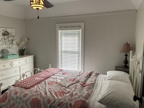 Une chambre à coucher comprenant un lit, une commode, une fenêtre avec des stores, un ventilateur de plafond et une lampe sur une table de chevet.