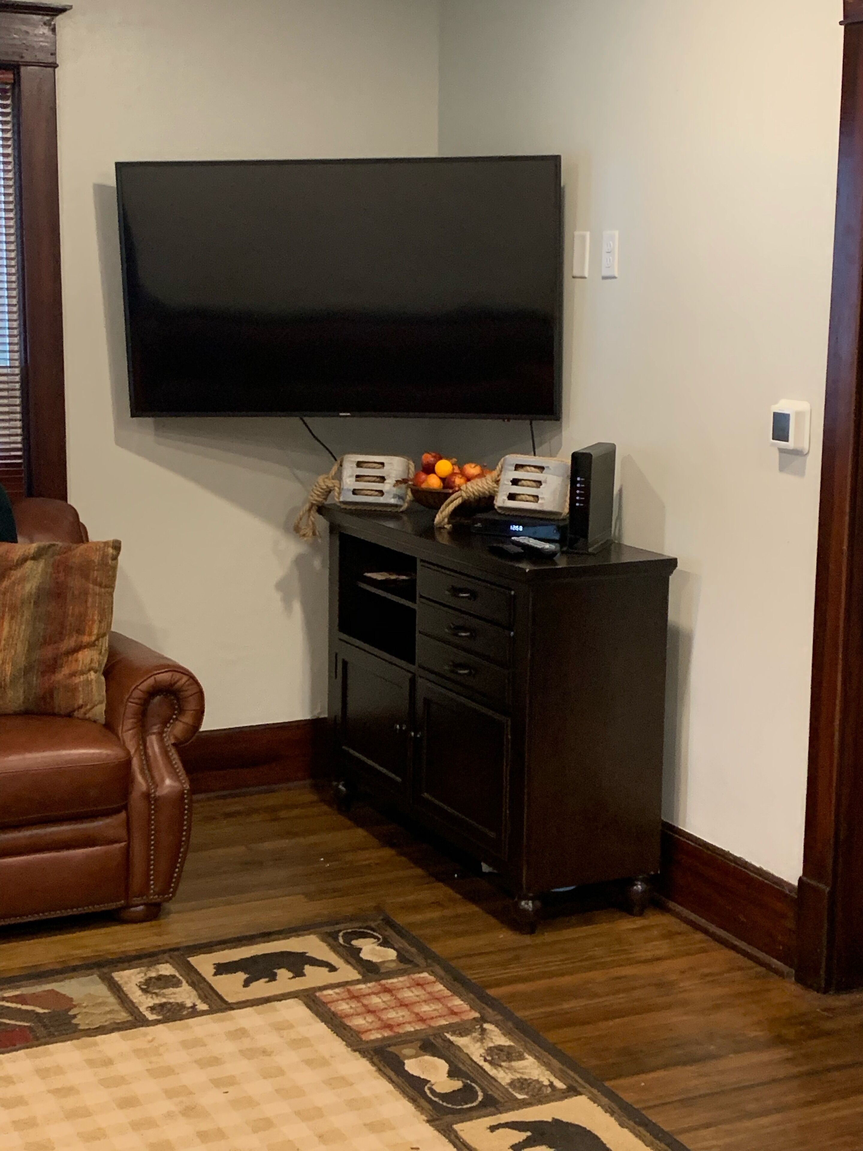 Un salon avec un téléviseur à écran plat fixé au mur, un meuble en bois foncé contenant un panier de fruits et des appareils électroniques, un canapé en cuir marron et un tapis à motifs.
