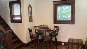 Zona pranzo con una scacchiera, sedie di legno, una scala e una finestra con inferriata decorativa.