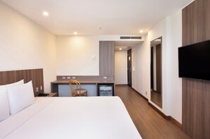 Quarto de hotel com cama, escrivaninha, cadeira, televisão e um pequeno frigorífico.