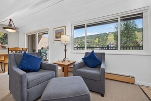 Une pièce avec deux fauteuils gris, une petite table en bois et une lampe. Une grande fenêtre offre une vue sur un paysage verdoyant.
