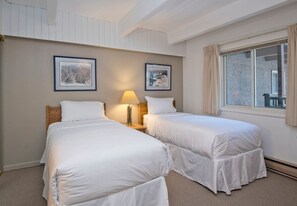 A hotel room with two single beds, a nightstand with a lamp, framed pictures on the wall, and a window with curtains.