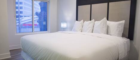 Una habitación de hotel moderna con una cama grande, un ventanal con vistas a la ciudad y una mesita de noche sencilla con una lámpara.