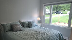 Un dormitorio con cama, almohadas, una mesita de noche, lámpara y ventana con vista a la calle y vegetación.