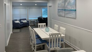 Un moderno comedor con una mesa y sillas blancas, un centro de mesa de vidrio azul y vista a una sala de estar con un sofá y un cuadro.
