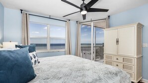 Une chambre à coucher avec un lit, un ventilateur de plafond, une grande fenêtre donnant sur l’océan et une armoire blanche.