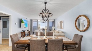 Un espace repas comprenant une table en bois, des chaises rembourrées, un lustre et une vue sur l’océan grâce à de grandes fenêtres.