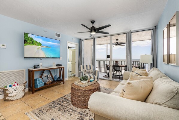 A living room with a large TV, a ceiling fan, a wicker ottoman, and a sofa. - Remodeled Gulf-Front Condo | 3rd Floor 180° Views & Walk to Times Square (Fort Myers Beach)