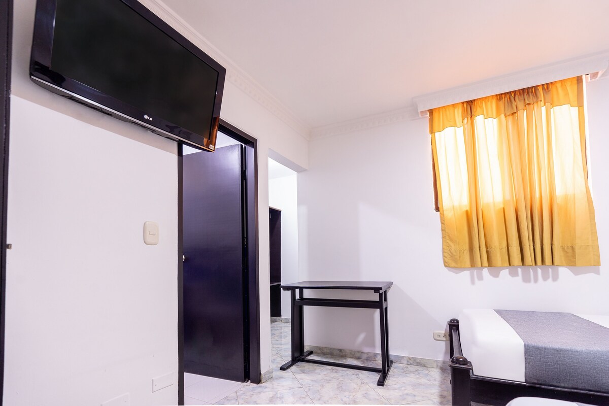 A room with a bed, a TV mounted on the wall, a table, and a window with curtains.