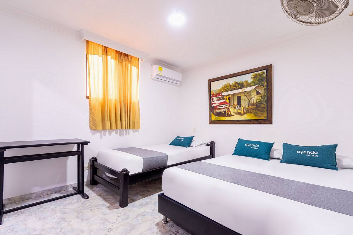 A hotel room with two single beds, a desk, a window with curtains, an air conditioning unit, and a painting on the wall.