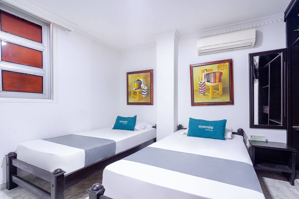 Two single beds with grey and white bedding, a small bedside table, and a wall-mounted air conditioner.