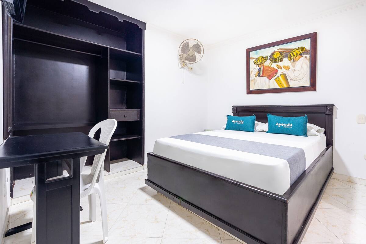 A bedroom with a bed, a desk, a chair, a wall-mounted fan, and a painting.