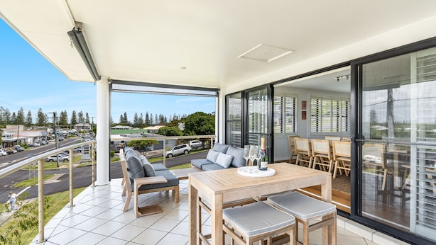 A modern balcony with a wooden table and chairs, a sofa, and a view of a street and greenery.