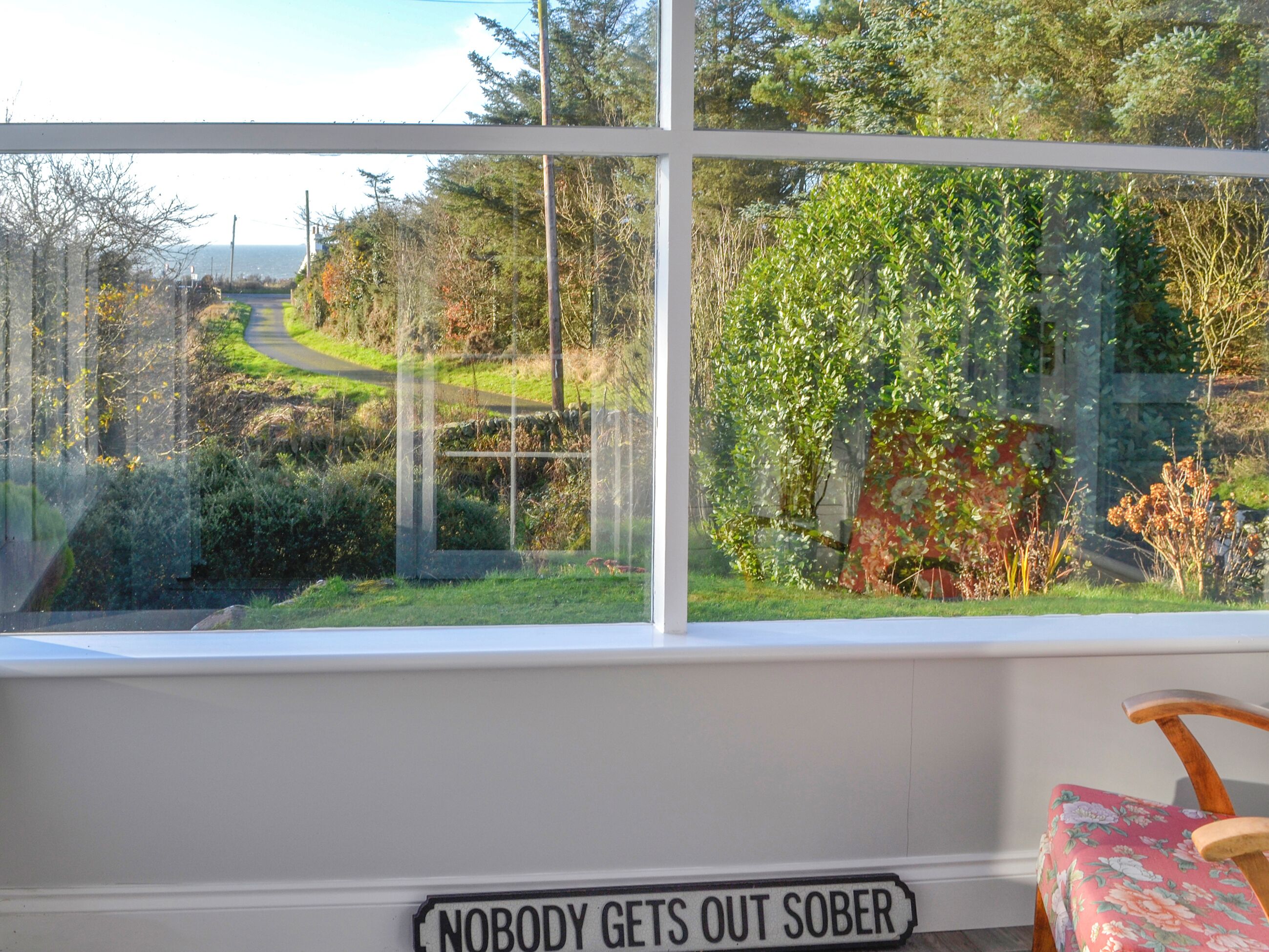 Ein Zimmer mit einem großen Fenster, das einen Blick auf eine kurvenreiche Straße und Grünflächen bietet. Auf dem Fenster steht ein Schild mit der Aufschrift 'NIEMAND KOMMT HIER SÖBER RAUS.'