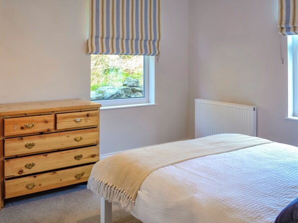 Een slaapkamer met een houten dressoir, een bed met een beige deken, een raam met gestreepte gordijnen en een radiator.
