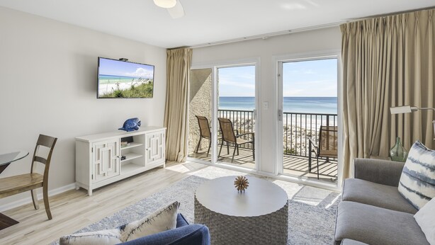 A modern living room with a television, a sofa, a coffee table, and a balcony with a view of the beach.