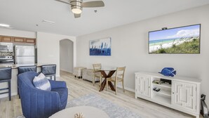 A modern living room with a dining area, a ceiling fan, a flat-screen TV, and a kitchen in the background.