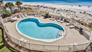 A pool with a heart-shaped design, surrounded by a white fence and lounge chairs, overlooking a beach with sand dunes and the ocean.
