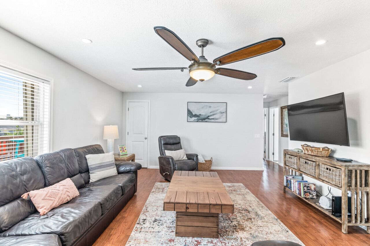 A modern living room with a leather sofa, a wooden coffee table, a black leather armchair, a ceiling fan, a TV, and a bookshelf.
