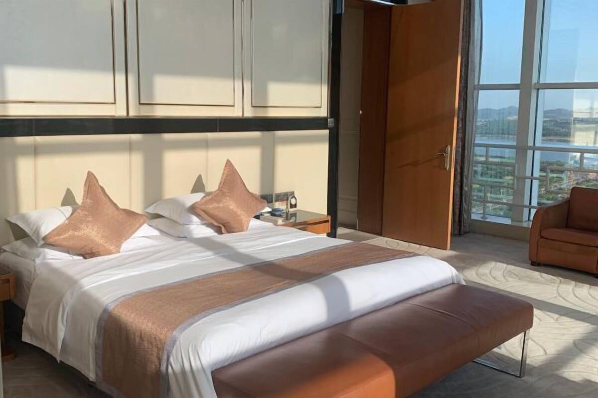 Habitación de hotel con una cama grande, un banco de madera, una mesita de noche y vistas amplias a través de un ventanal.